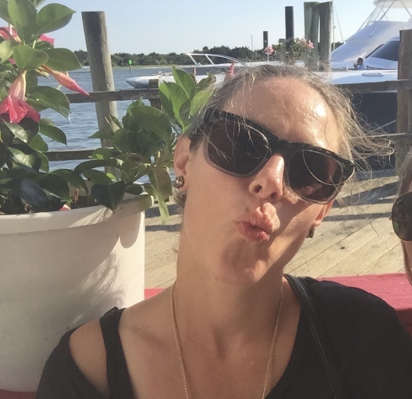 photo of Staci (light-skinned woman) wearing dark sunglasses and making a kissy face in front of the ocean