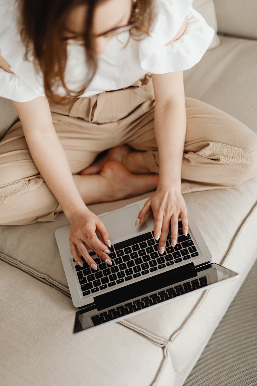 a light-skinned woman sitting cross legged on a sofa and typing on a computer